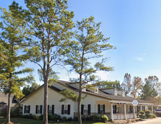 Single-story white senior living building with dark shutters, a porch, and tall trees.