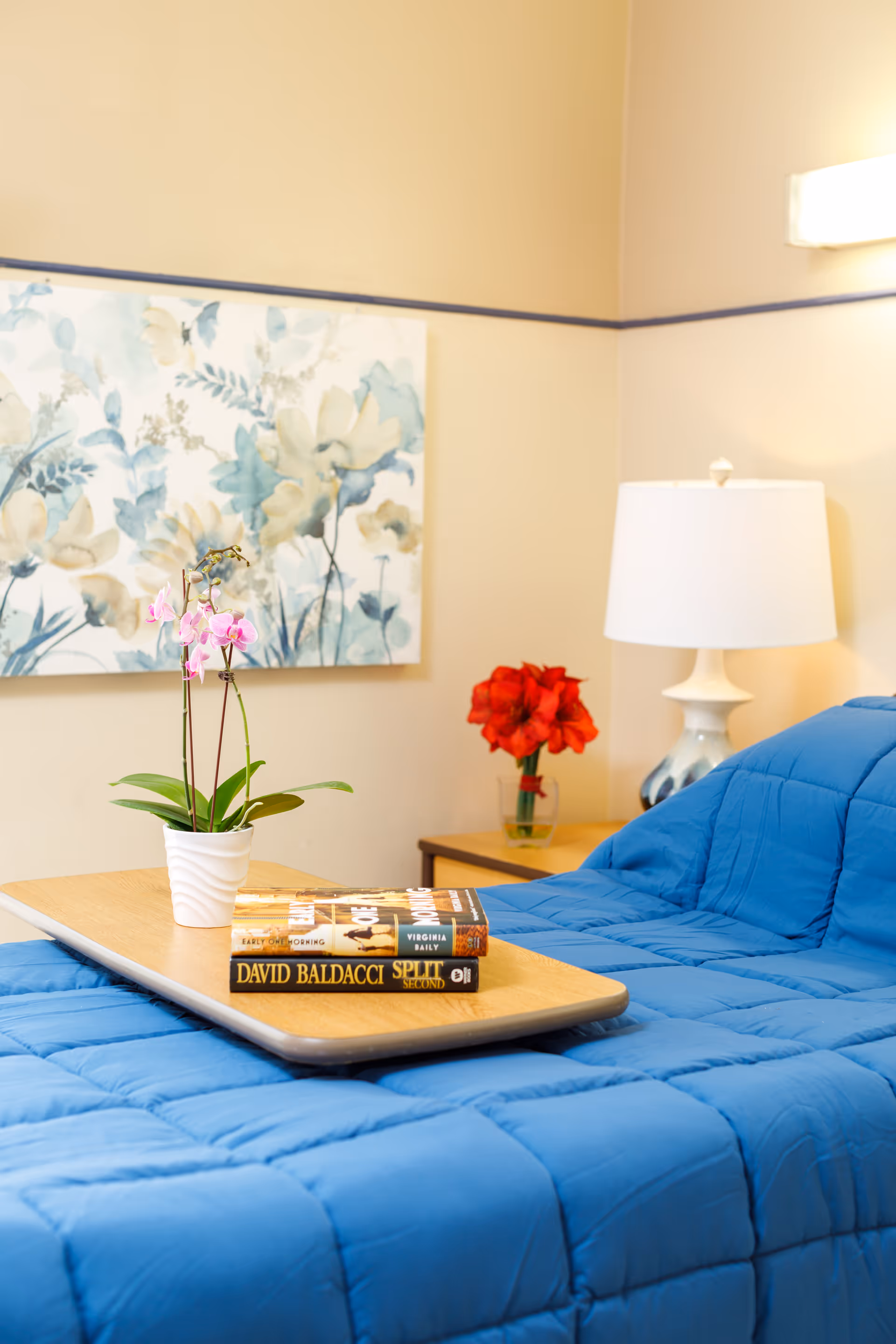A cozy bedroom with a blue quilted bedspread, a wooden tray on the bed holding a small potted orchid and two books. In the background, there is a nightstand with a white lamp and a vase with red flowers, and a floral painting on the beige wall.