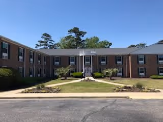Front exterior view of a two-story brick building with multiple windows and a central entrance with white columns, surrounded by a well-maintained lawn and small shrubs under a clear blue sky.