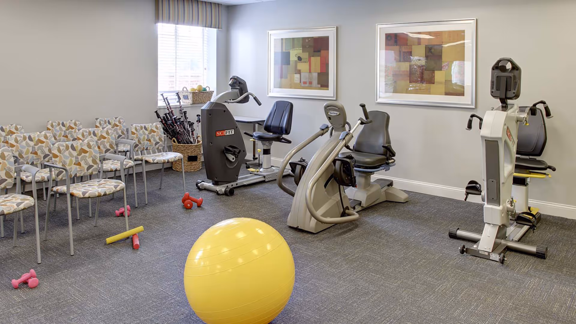 A small exercise room with three recumbent exercise bikes, a large yellow exercise ball, small dumbbells, and several chairs with patterned upholstery arranged along one wall. Two framed abstract artworks hang on the wall above the exercise bikes, and a window with blinds lets in natural light.