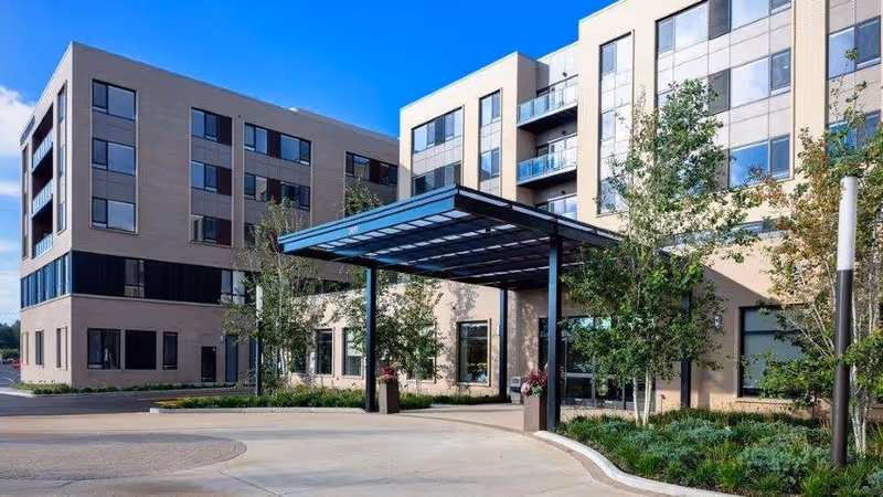 Exterior view of a modern multi-story senior living facility with large windows, a covered entrance, and landscaped greenery under a clear blue sky.