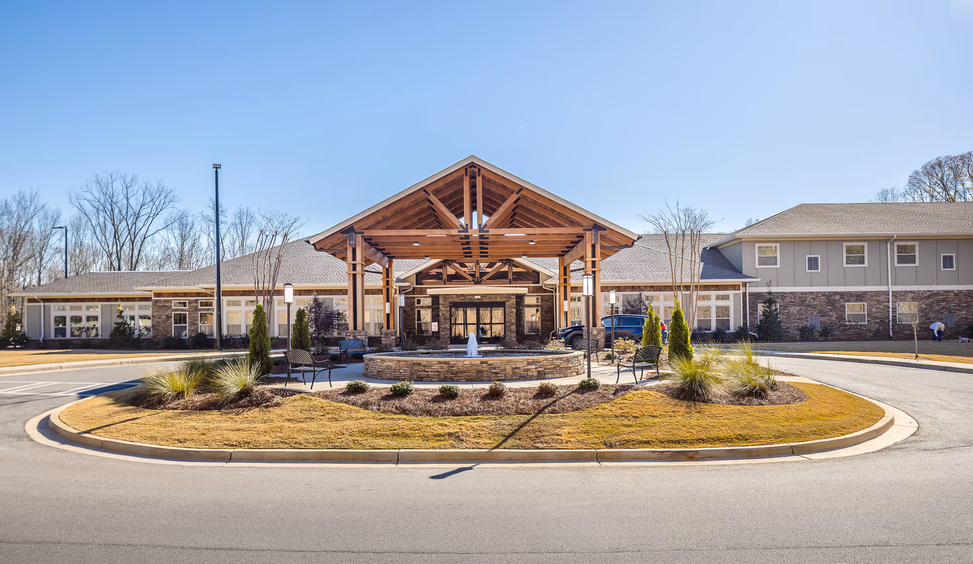 Front entrance of a large senior living building with a timber-covered porte-cochere, circular driveway, fountain, benches and landscaped island.