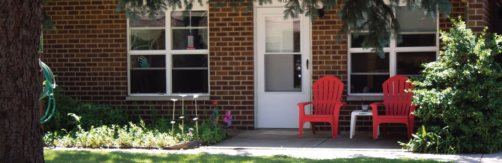 Outdoor patio area of a brick building with two red Adirondack chairs and a small white table between them. There are two windows and a white door behind the chairs. Green bushes and plants surround the patio, and a tree trunk is visible on the left side of the image.