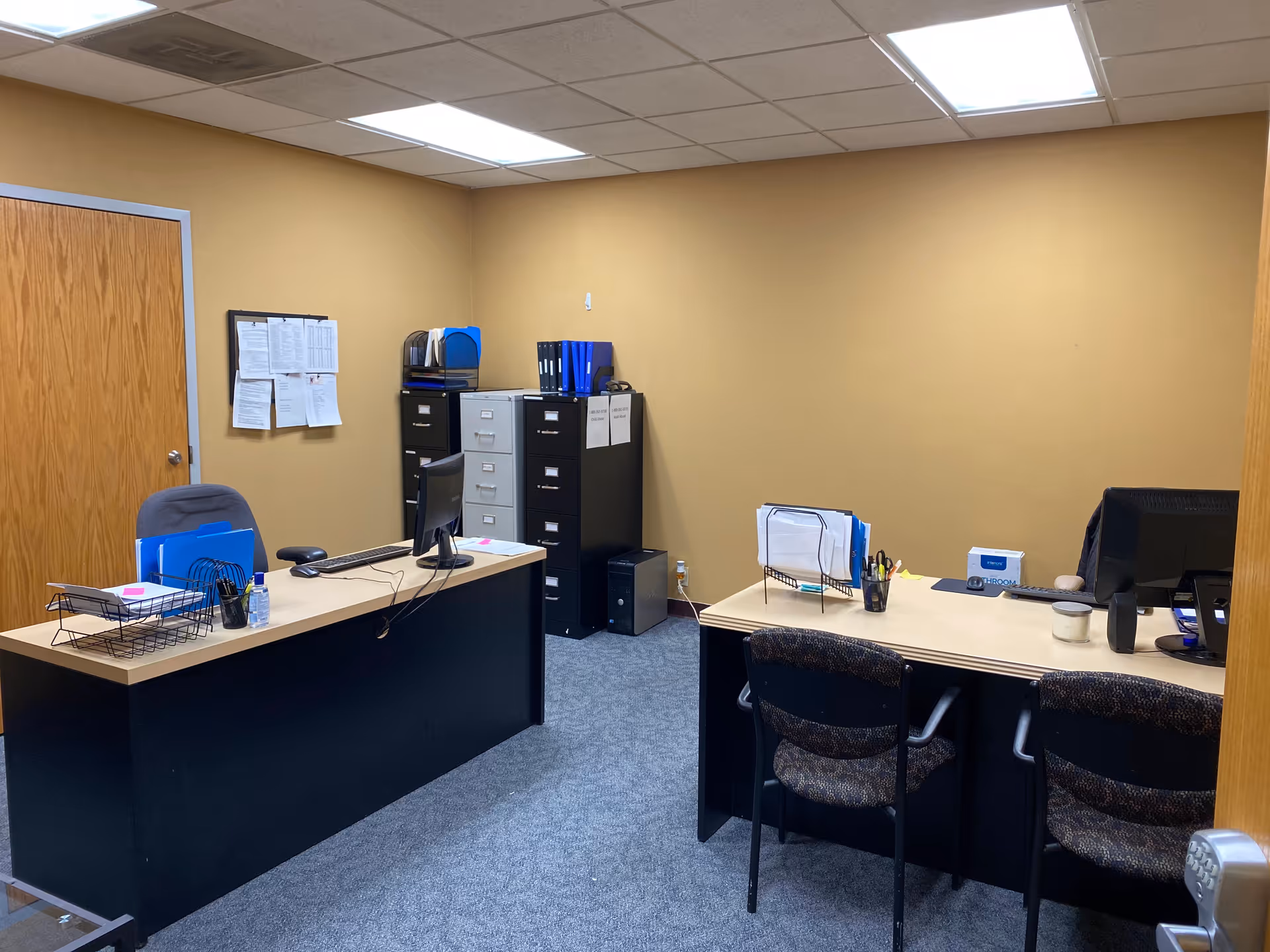 Office workspace with two desks, filing cabinets, and office chairs in a beige-walled room.