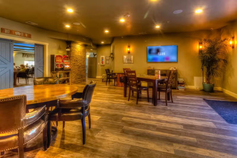 Interior view of a senior living facility common area with wooden tables and chairs, warm lighting, a flat-screen TV mounted on the wall, a potted plant in the corner, and a doorway leading to another room with more seating visible.
