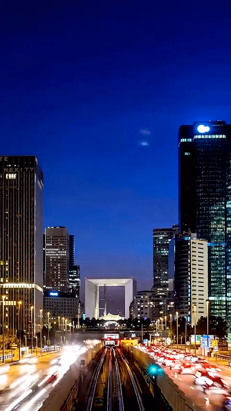 Nighttime cityscape with tall buildings and light-trail traffic along a multi-lane road.