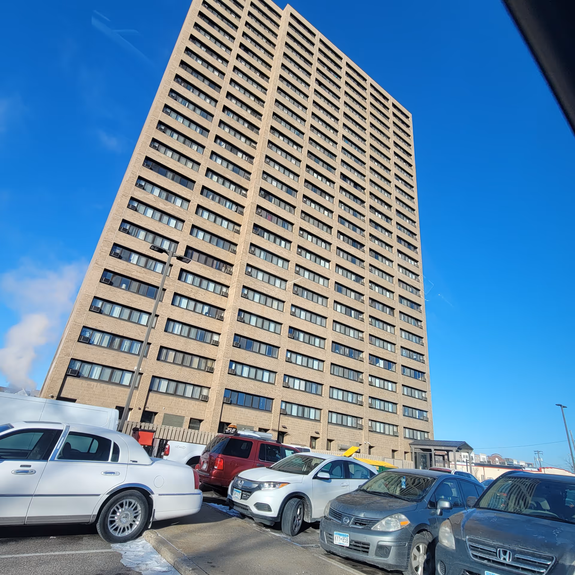 Tall beige high-rise building with many windows looms over a row of parked cars.