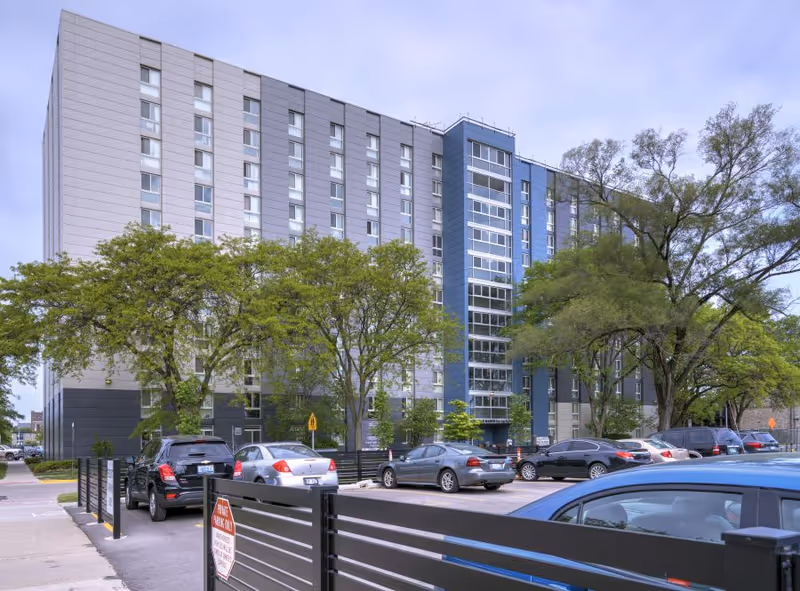 Exterior view of a multi-story residential building with trees and a parking lot in front.