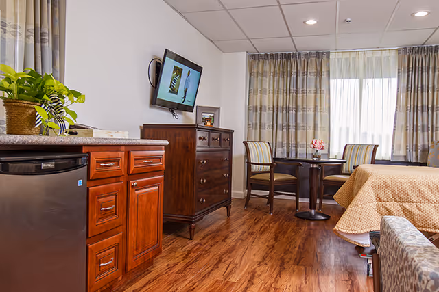 A cozy senior living suite with a bed, dresser, small table, TV, and kitchenette.
