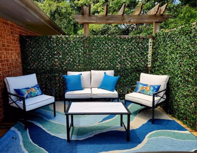 Outdoor seating area with a white cushioned loveseat and two matching armchairs, each with blue decorative pillows. A rectangular white coffee table with a black frame sits on a blue and green patterned outdoor rug. The area is enclosed by green leafy privacy screens and a wooden pergola overhead.