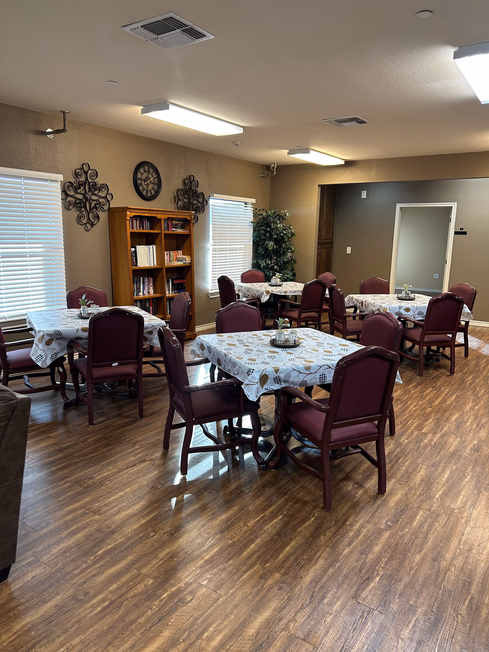 A dining room with round tables, maroon chairs, and a bookshelf against beige walls.