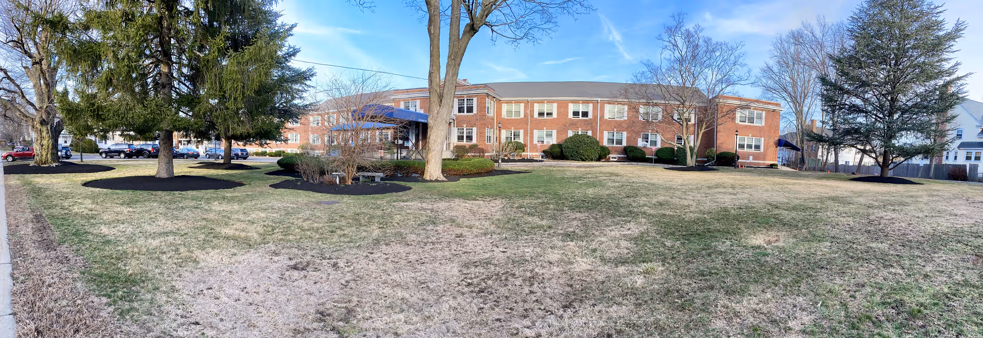 Brick senior care facility building with a lawn, trees, and parked cars.