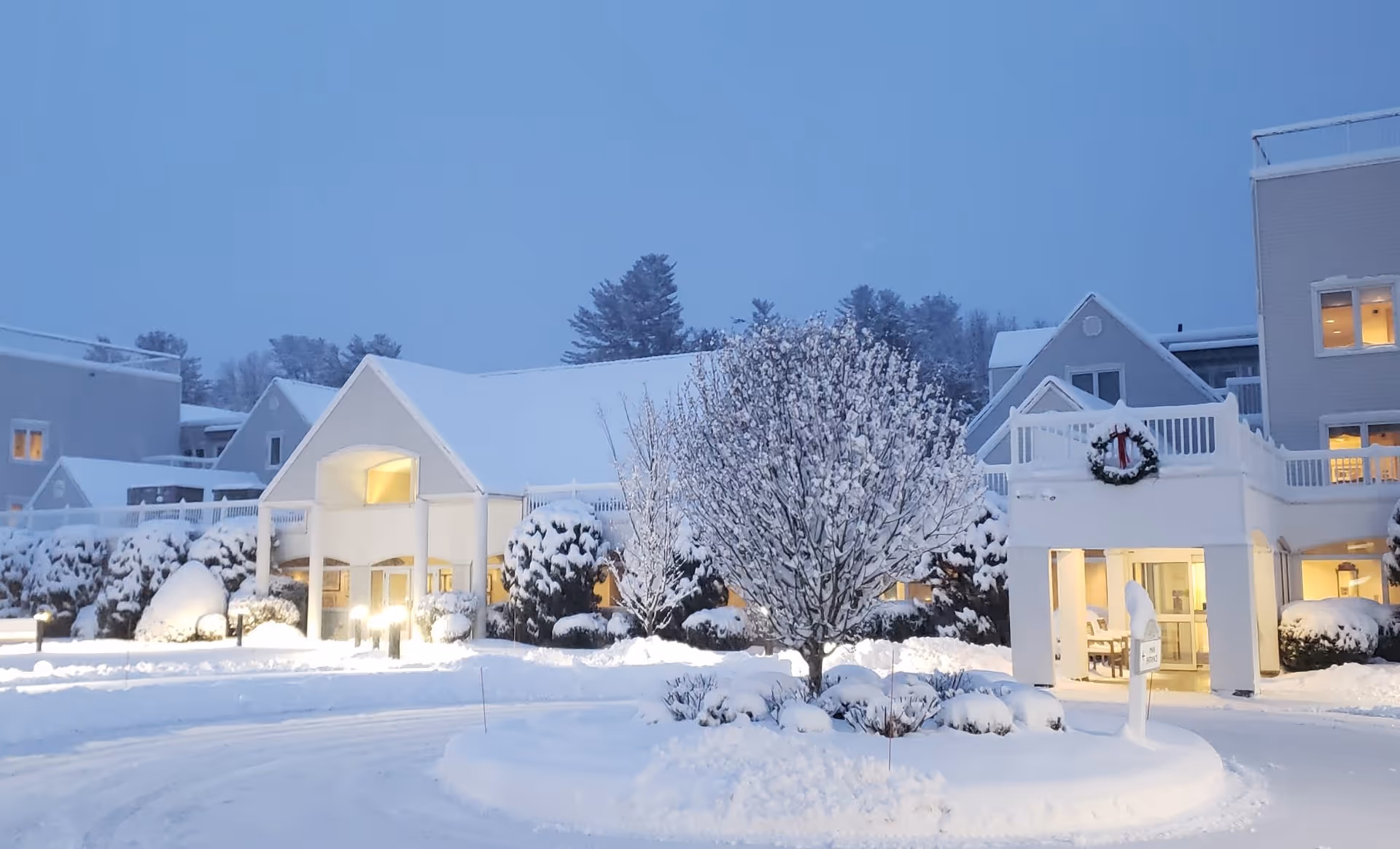 Snow-covered entrance of a senior living facility named Pomperaug Woods during winter evening, with lit windows and a decorated wreath on the building.