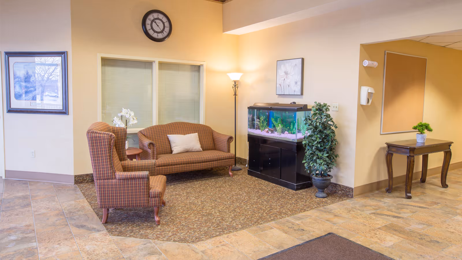 An indoor lounge area with plaid armchairs, a sofa, a fish tank, a tall floor lamp, framed art, and a small table with a plant.