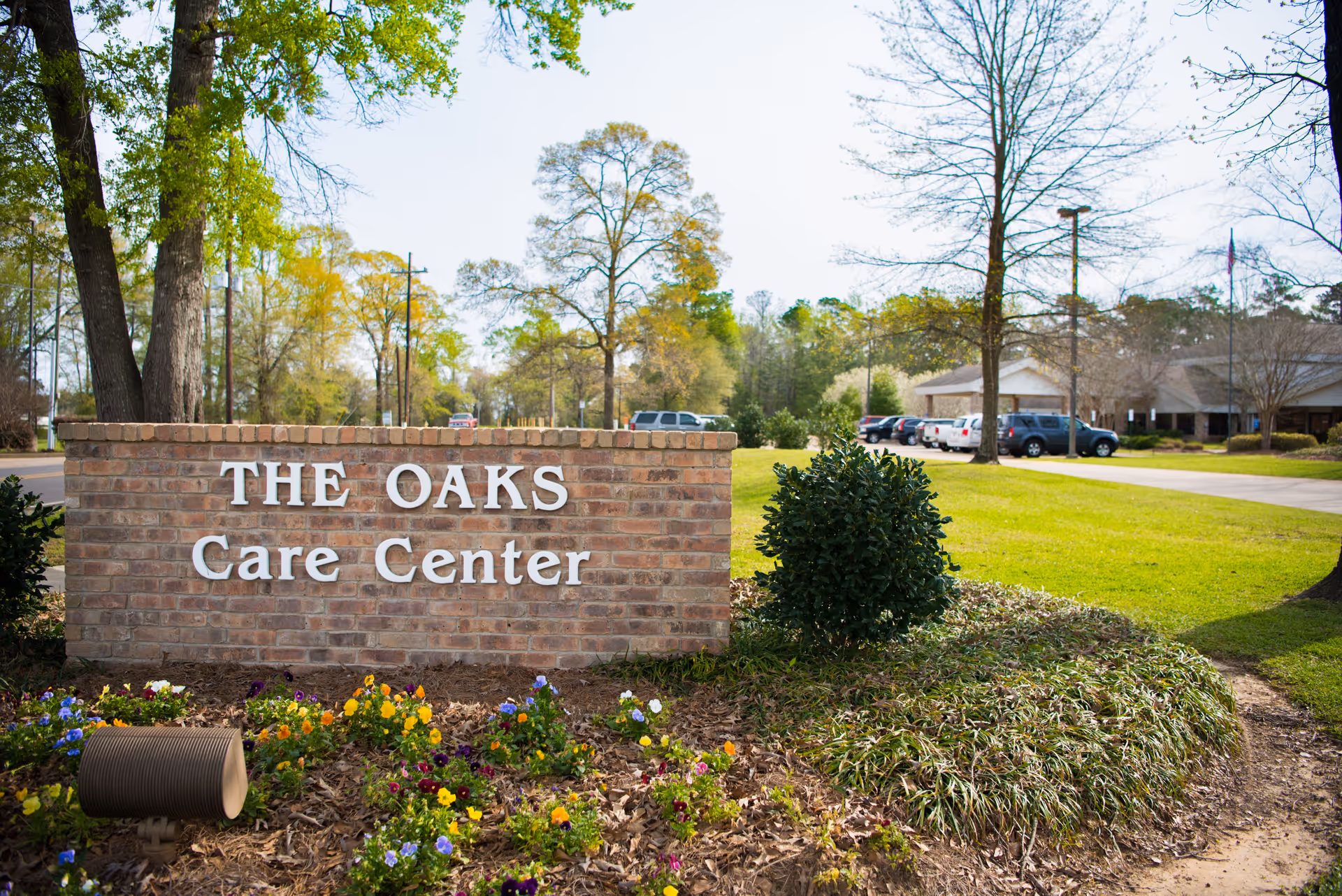 A brick sign with white letters that reads 'THE OAKS Care Center' surrounded by a landscaped area with flowers and bushes. In the background, there is a parking lot with several cars and a building partially visible among trees under a clear sky.