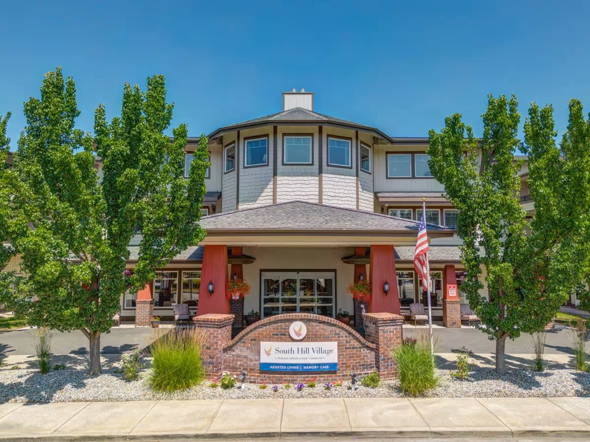 Front exterior view of South Hill Village senior living facility with a brick sign, American flag, trees, and a clear blue sky.