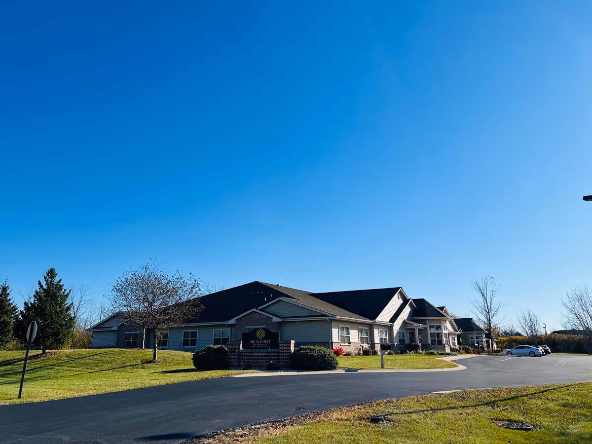 Front exterior of a single-story assisted living building with a circular driveway, lawn, and clear blue sky.