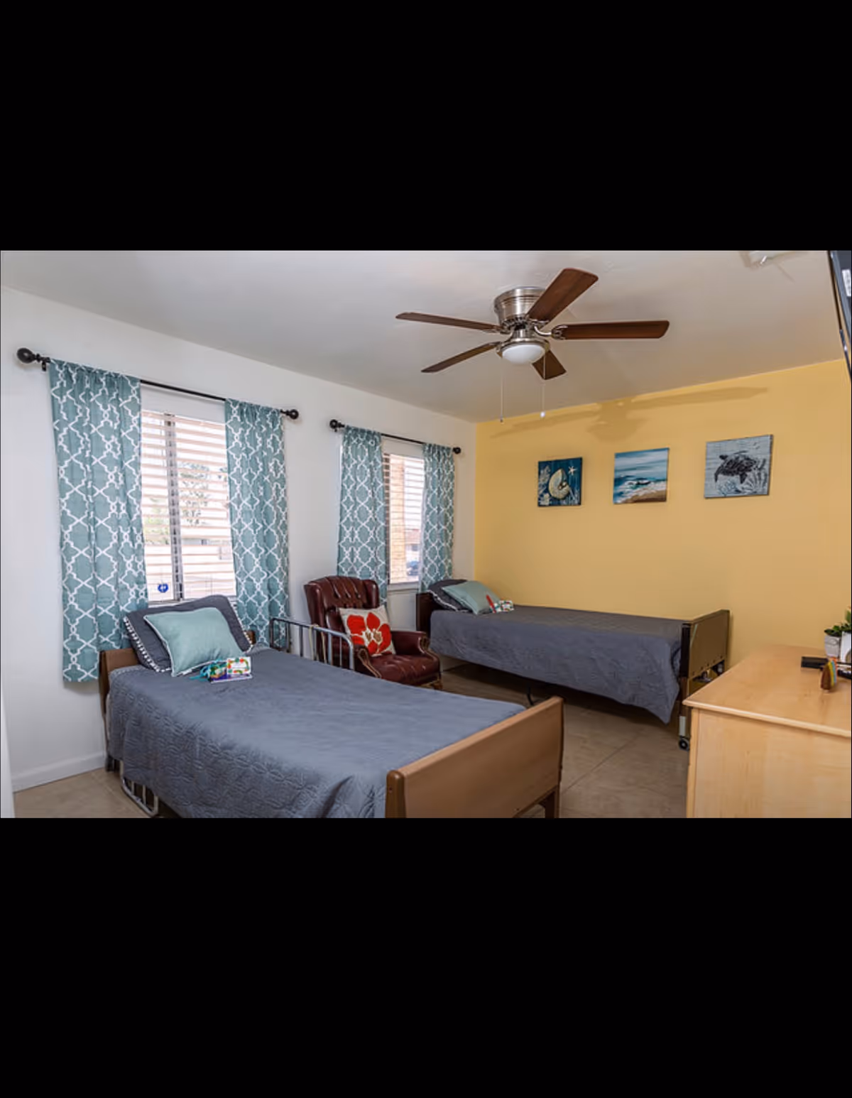 Twin bedroom with two beds, teal patterned curtains, a ceiling fan, and a yellow accent wall with artwork.