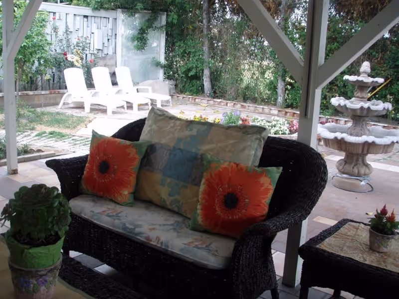 Outdoor covered patio area with a dark wicker loveseat featuring floral and patterned cushions, a small wicker table with a potted plant, a multi-tiered stone fountain, and three white lounge chairs in the background near a garden with flowers and trees.