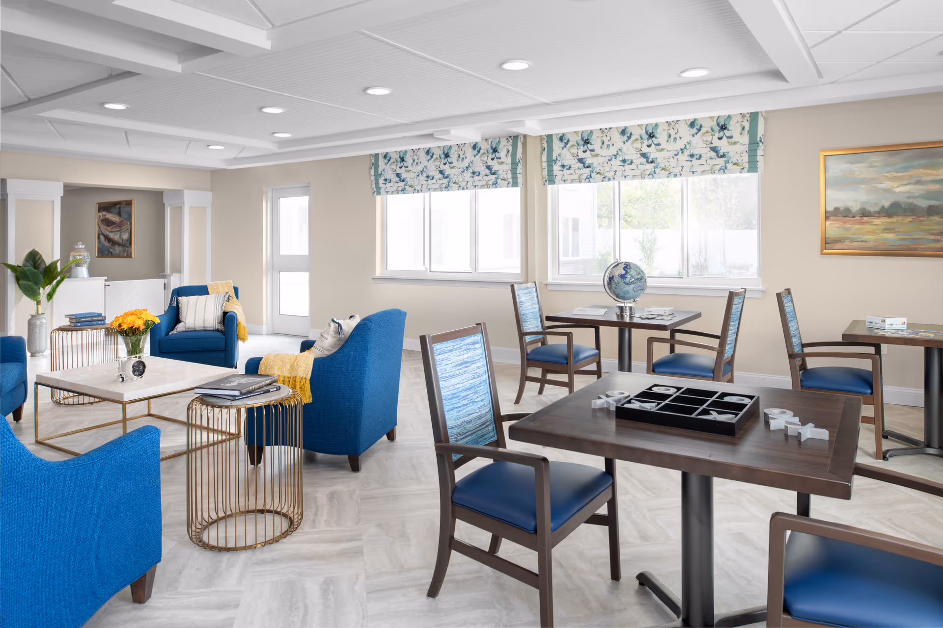 Bright communal lounge in a senior living facility with blue armchairs, game tables, and large windows.