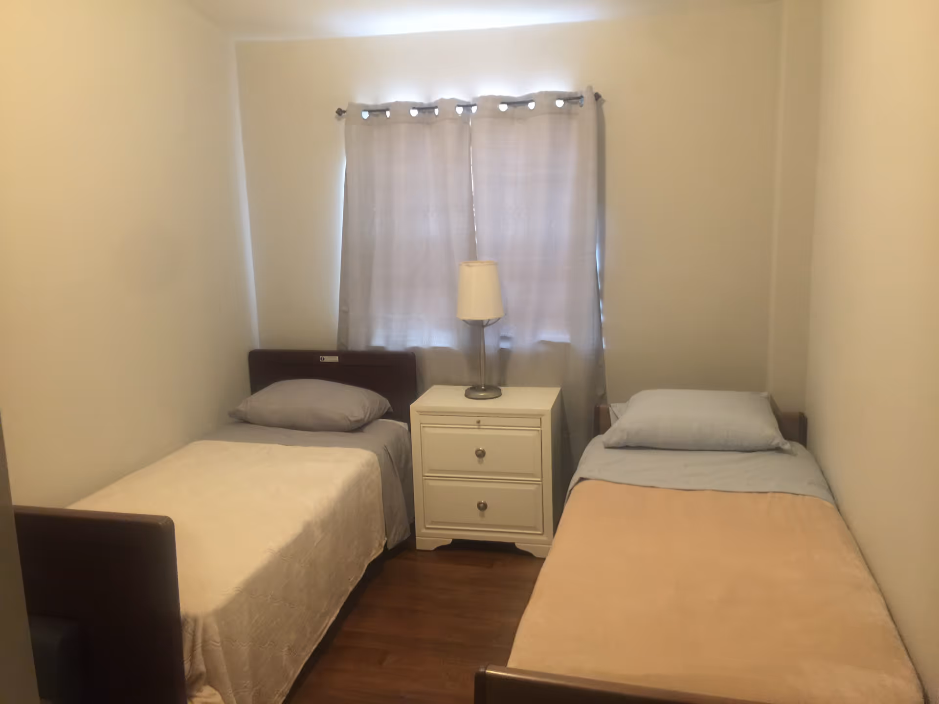 Two twin beds with a nightstand and lamp between, in a small senior living bedroom.