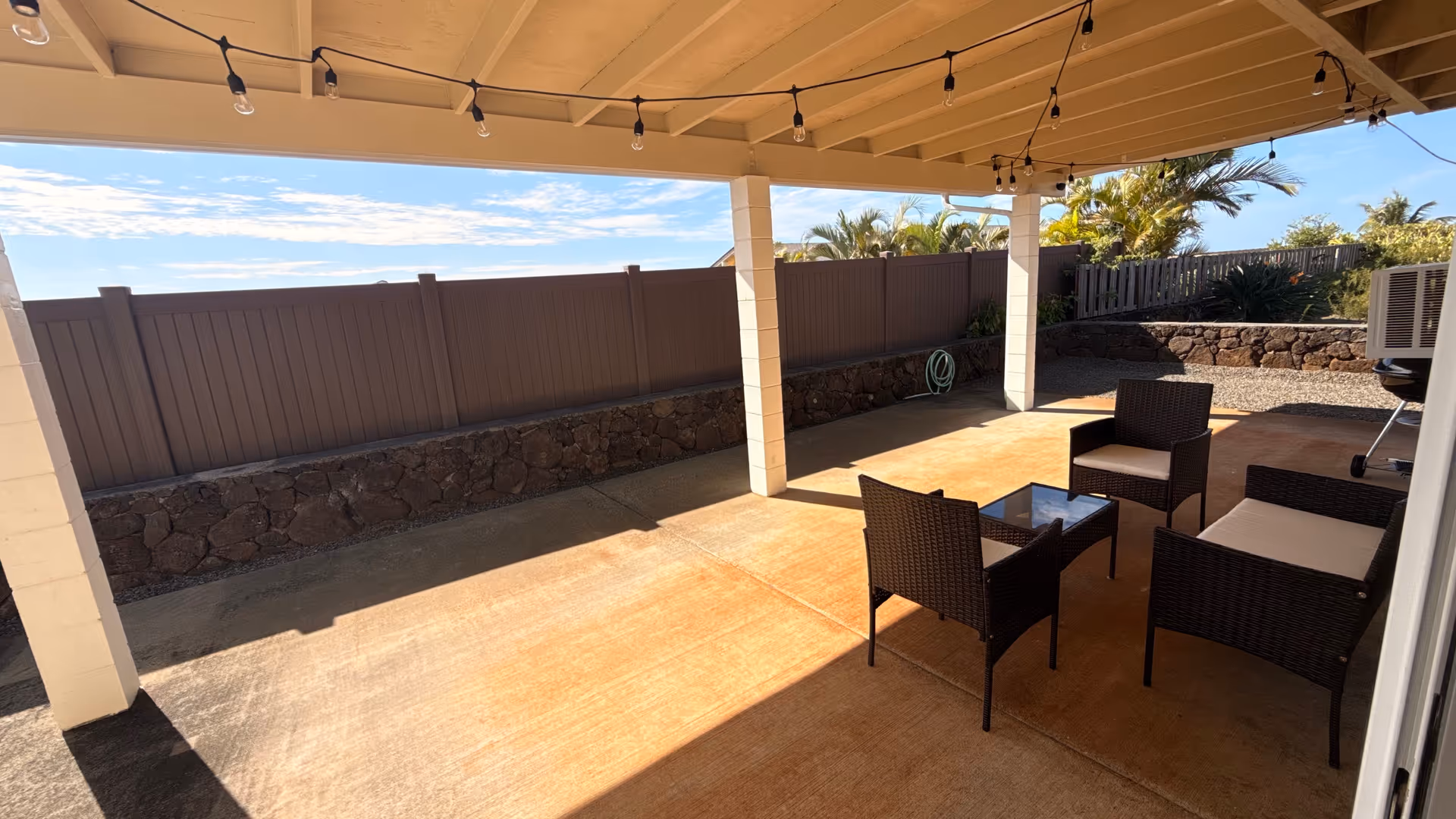 Covered patio with wicker seating, string lights, and a low stone wall.