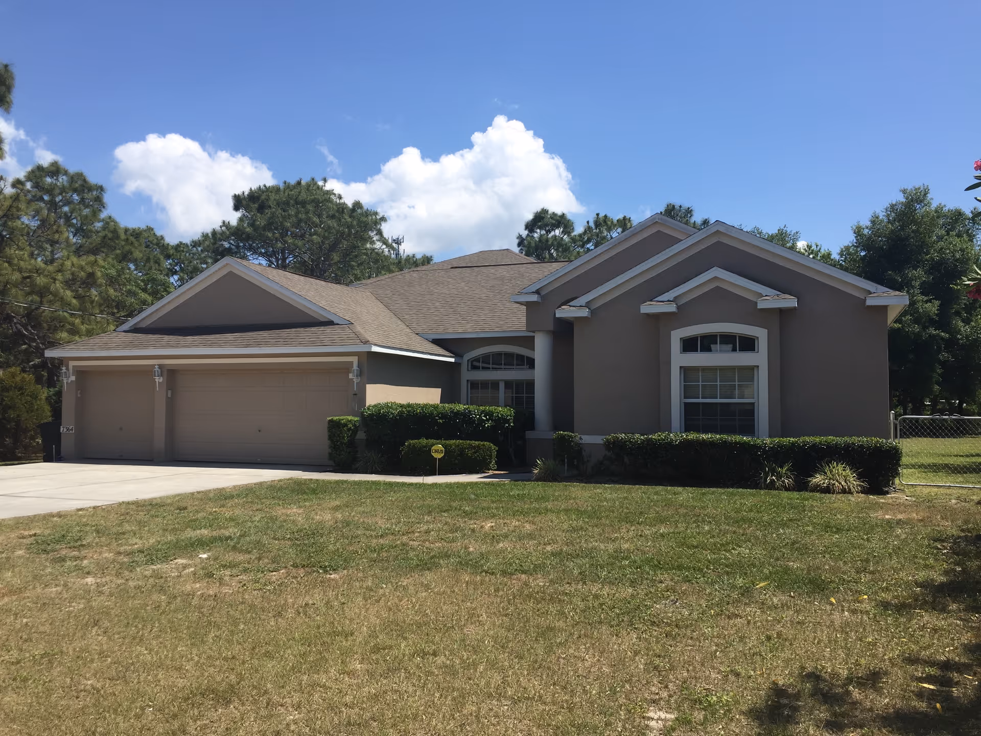 Single-story beige house with a three-car garage, trimmed shrubs and a front lawn under a blue sky.