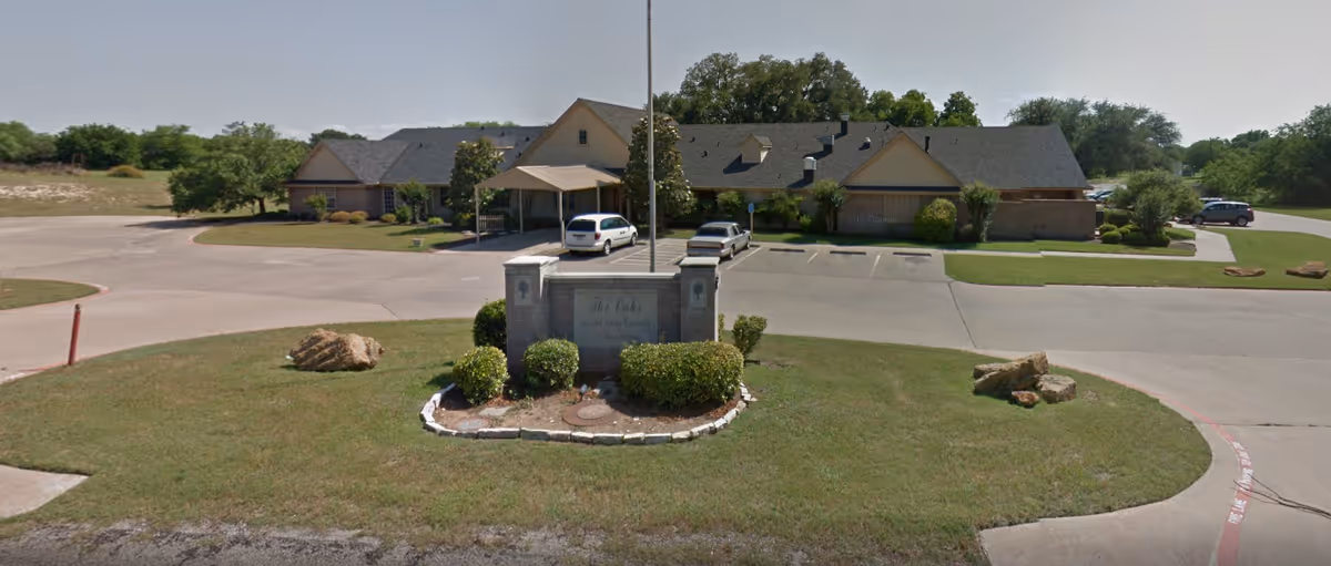 Front view of a single-story senior living building with an entrance sign, landscaped lawn, and parked cars in the driveway.