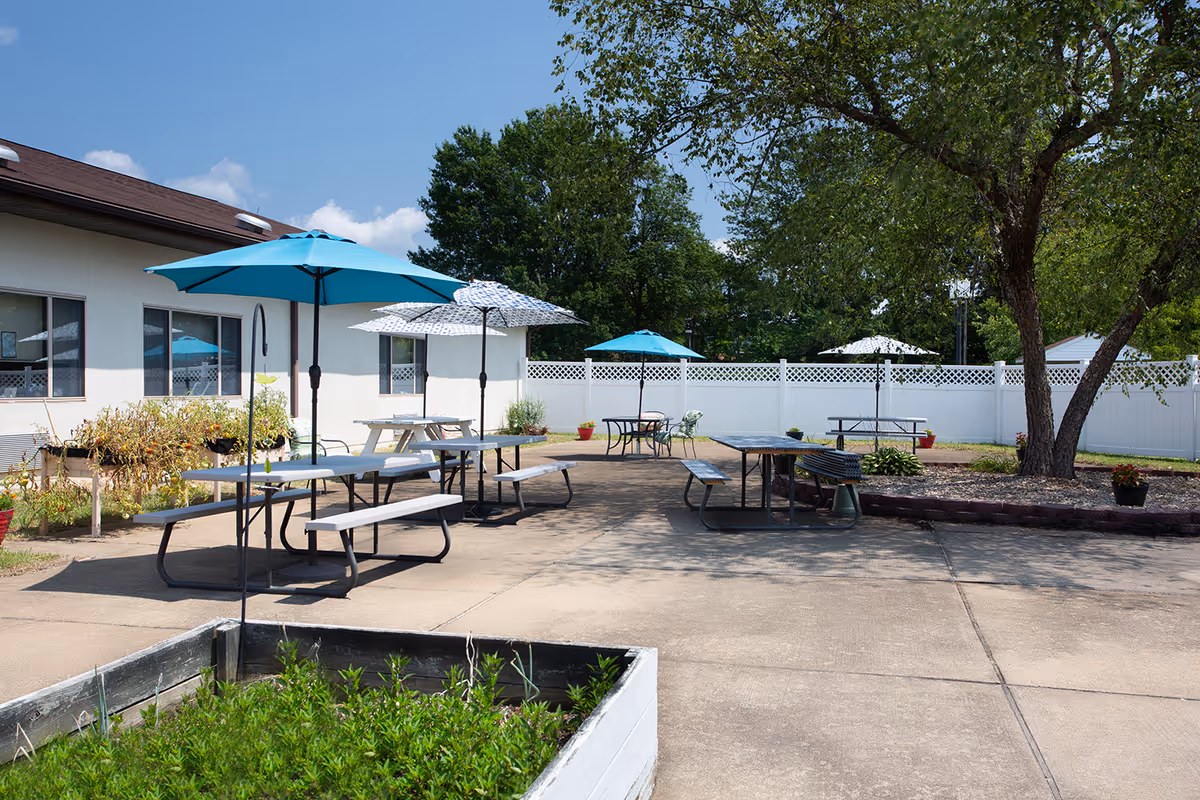 Outdoor courtyard with picnic tables under blue umbrellas beside a white fence and trees.