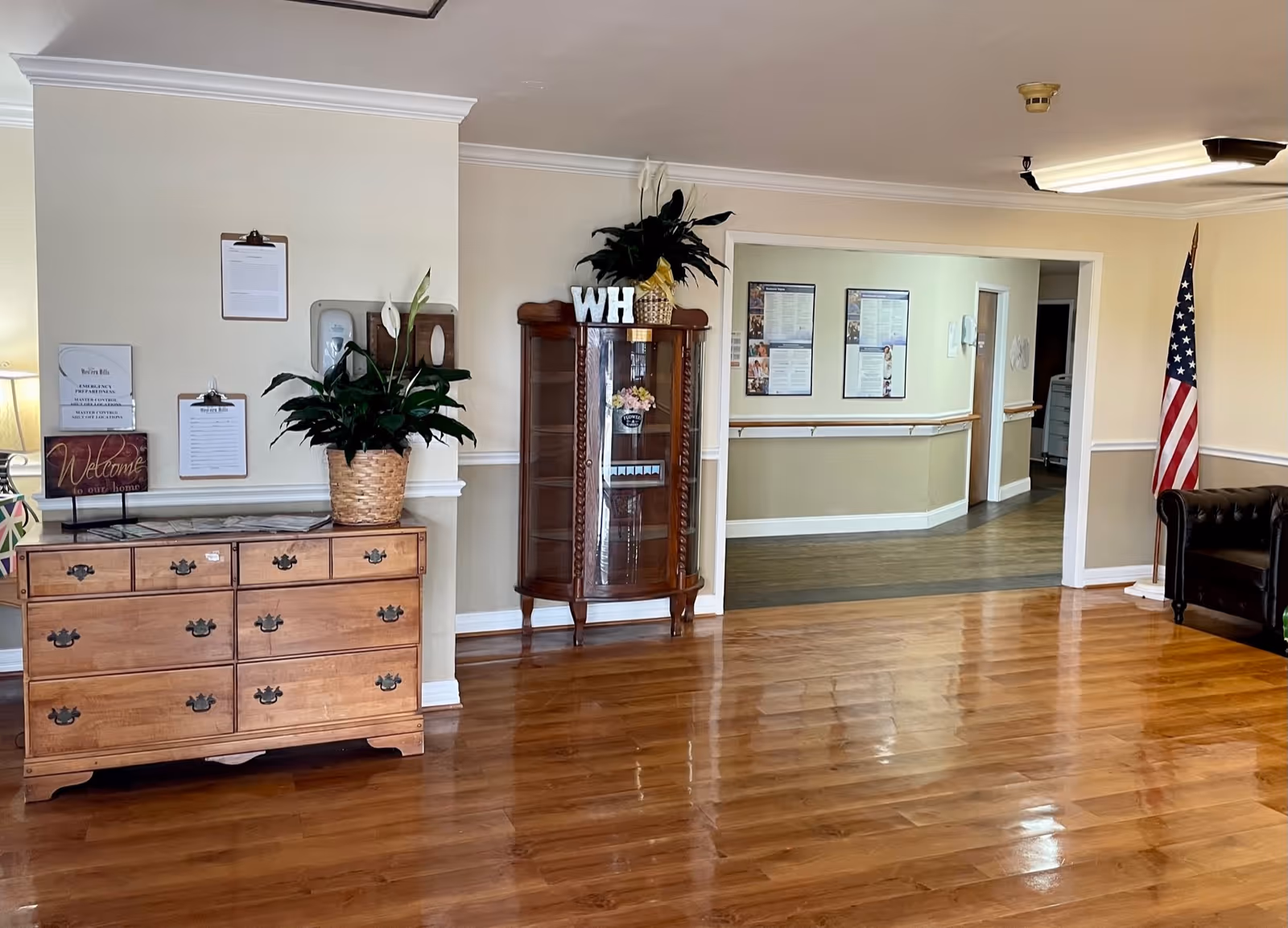 Interior view of a senior living facility with polished wooden floors, a wooden dresser with plants and clipboards on the wall, a glass display cabinet with decorative items and letters 'WH' on top, an American flag in the corner, and a hallway with handrails leading to other rooms.