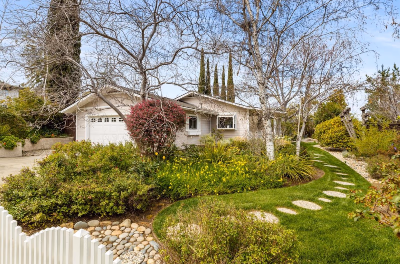 A single-story house with a white garage door surrounded by a lush garden. The garden features green grass, various bushes, flowering plants, and several leafless trees. A curved stone pathway leads through the garden, and a white picket fence is visible in the foreground under a clear blue sky.