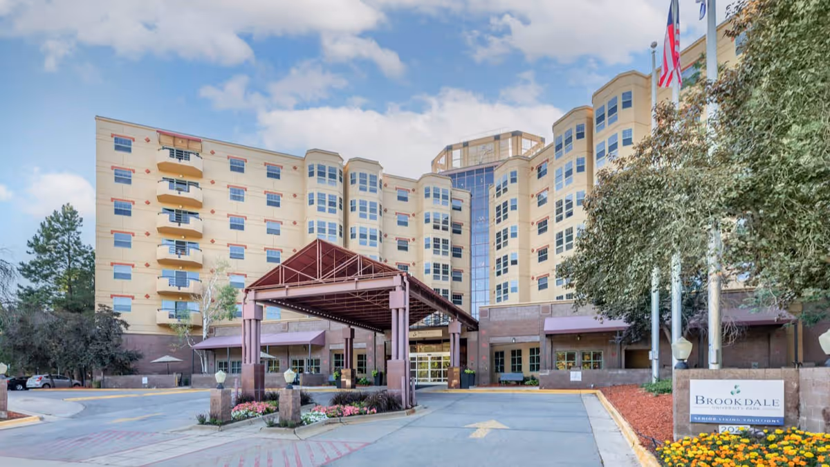 Front exterior view of Brookdale University Park, a multi-story senior living facility with a covered entrance, landscaped flower beds, and several flags near the entrance.