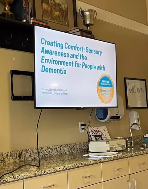 Wall-mounted presentation screen displaying dementia care seminar in a kitchen-like lounge area
