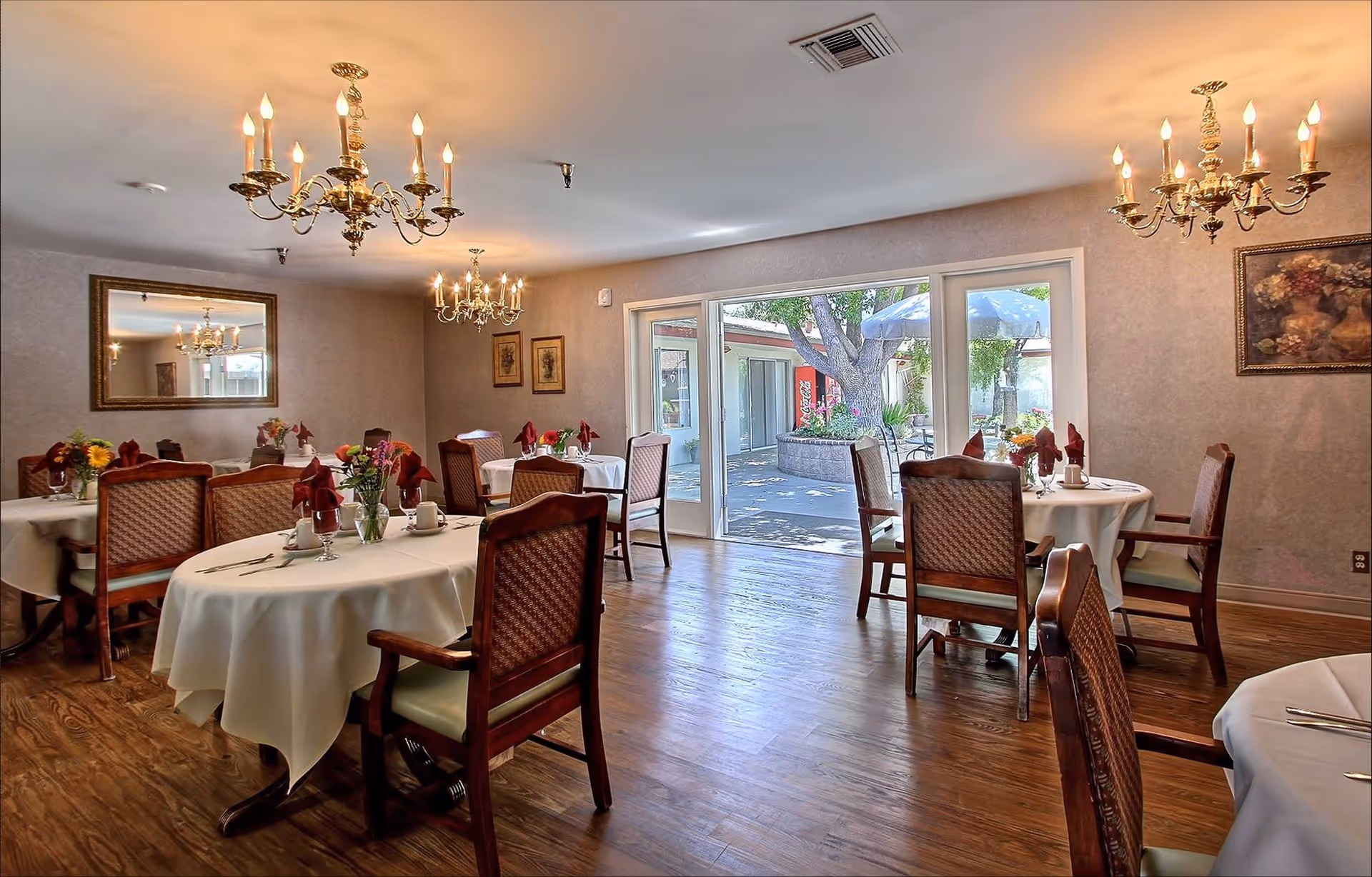 A dining room with round tables, white tablecloths, chandeliers, and glass doors to outdoors.