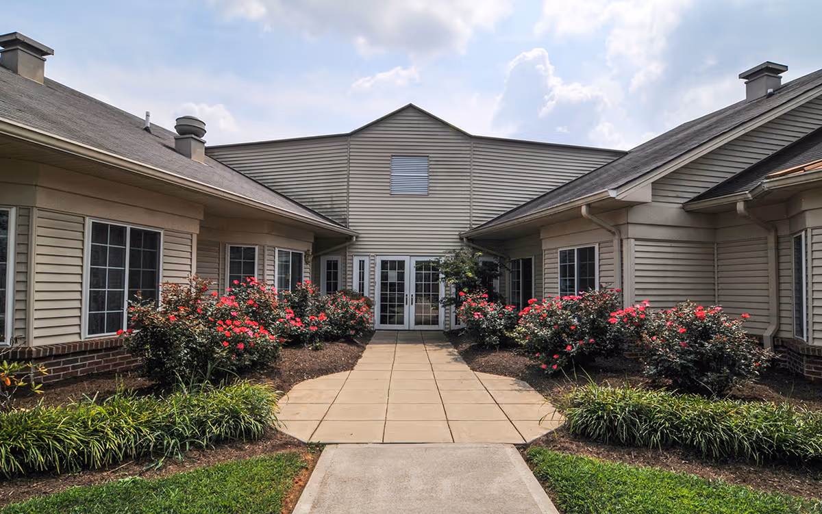Beige senior living facility exterior with a central entry and rose bushes lining a walkway.