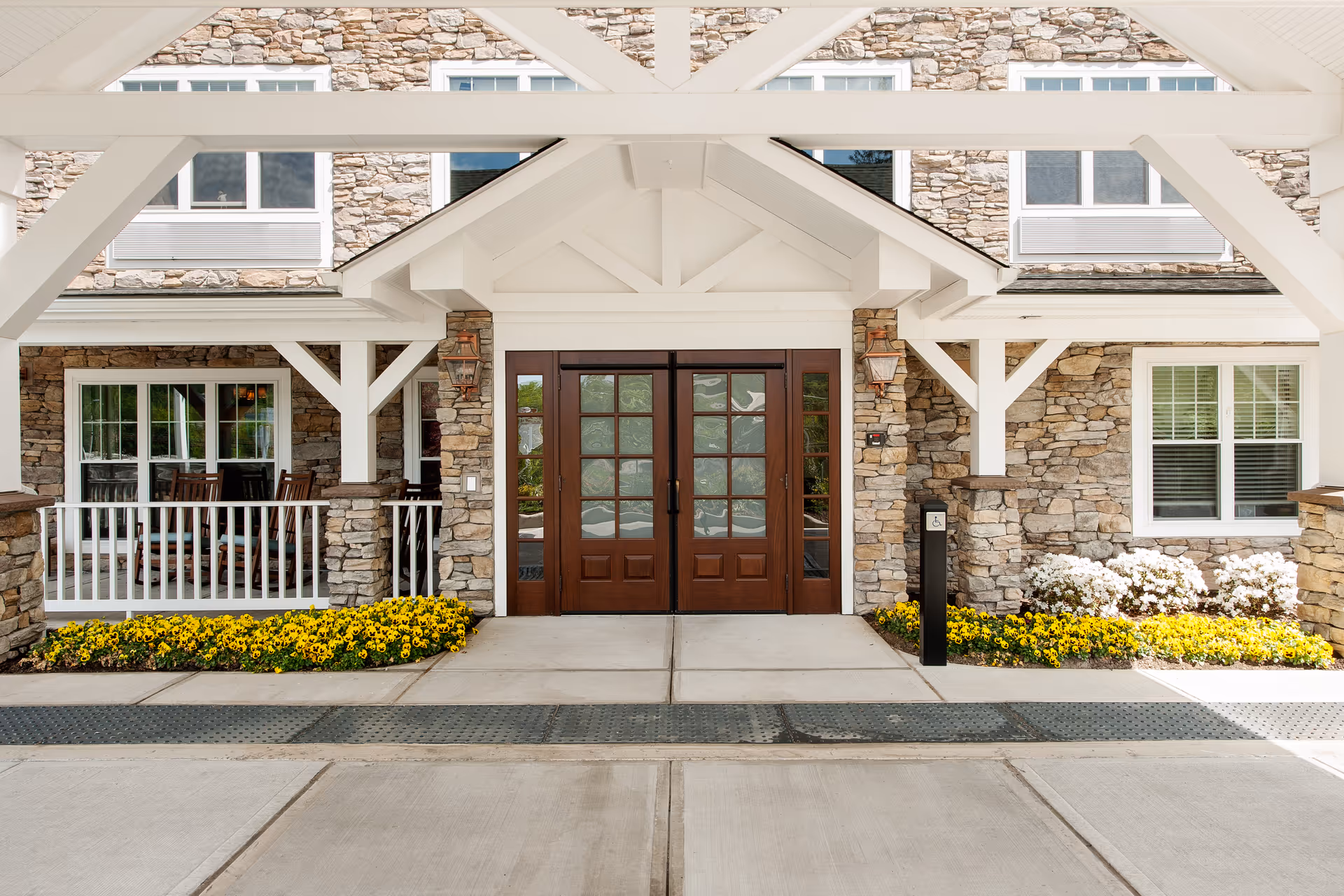 Entrance of a senior living facility with stone walls, white wooden beams, double wooden doors with glass panels, flower beds with yellow and white flowers on either side, and a covered porch area with a white railing and chairs.