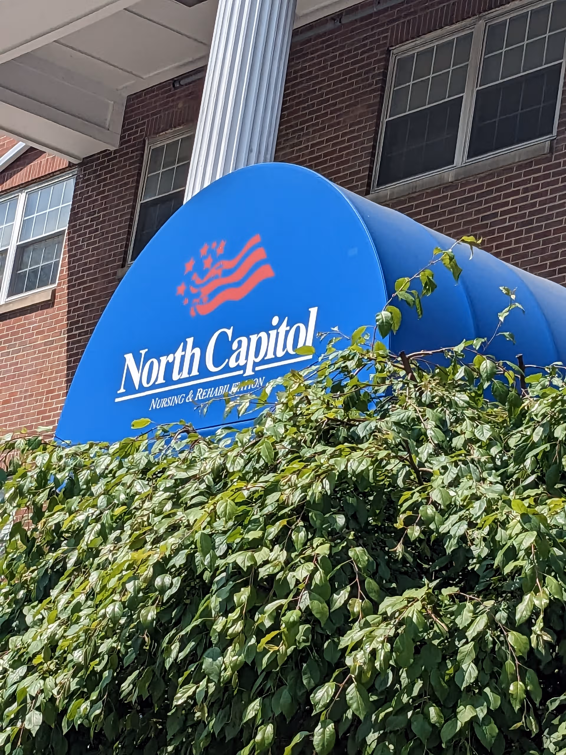 Blue curved building sign reading North Capitol Nursing and Rehabilitation, partially obscured by ivy.