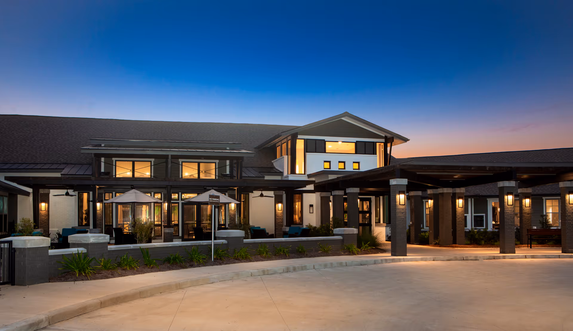 Exterior view of Avanti Senior Living at Lafayette during dusk, showing a modern building with large windows, outdoor seating areas with umbrellas, and a covered entrance with pillars and warm lighting.
