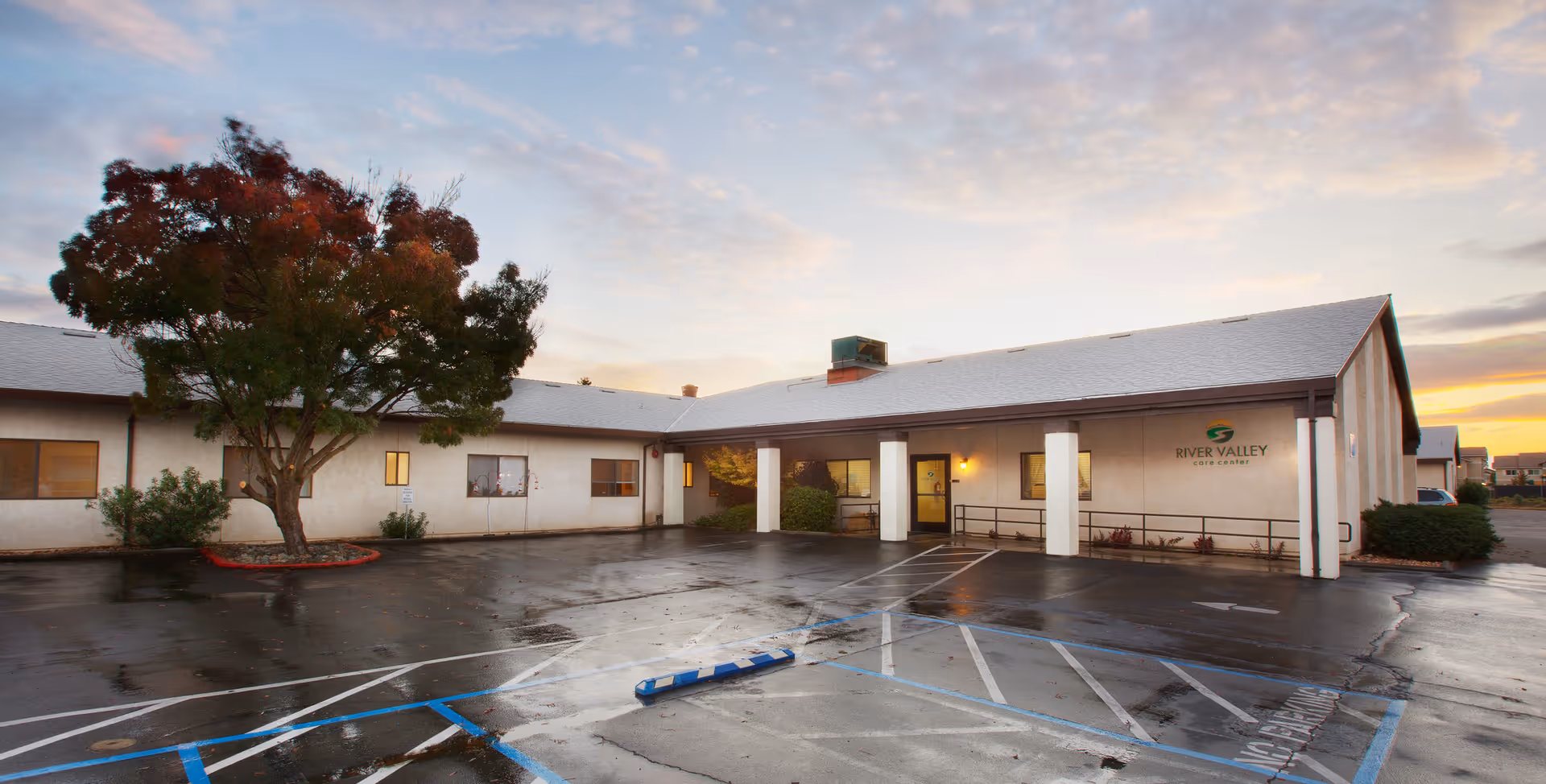 Exterior view of River Valley Care Center building during sunset with a large tree on the left and a wet parking lot with marked handicap spaces in front.