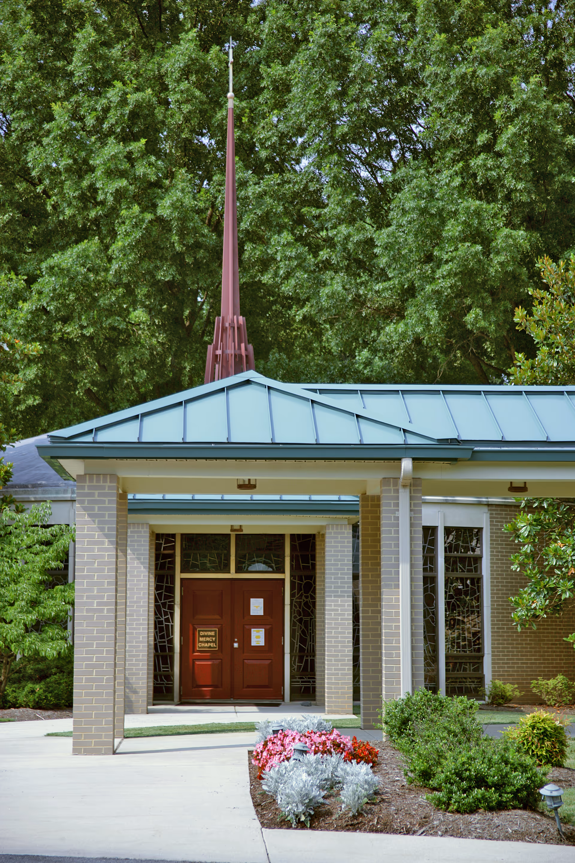 Entrance to a chapel building with a green metal roof and a tall red steeple, surrounded by lush green trees and landscaped bushes and flowers in front.
