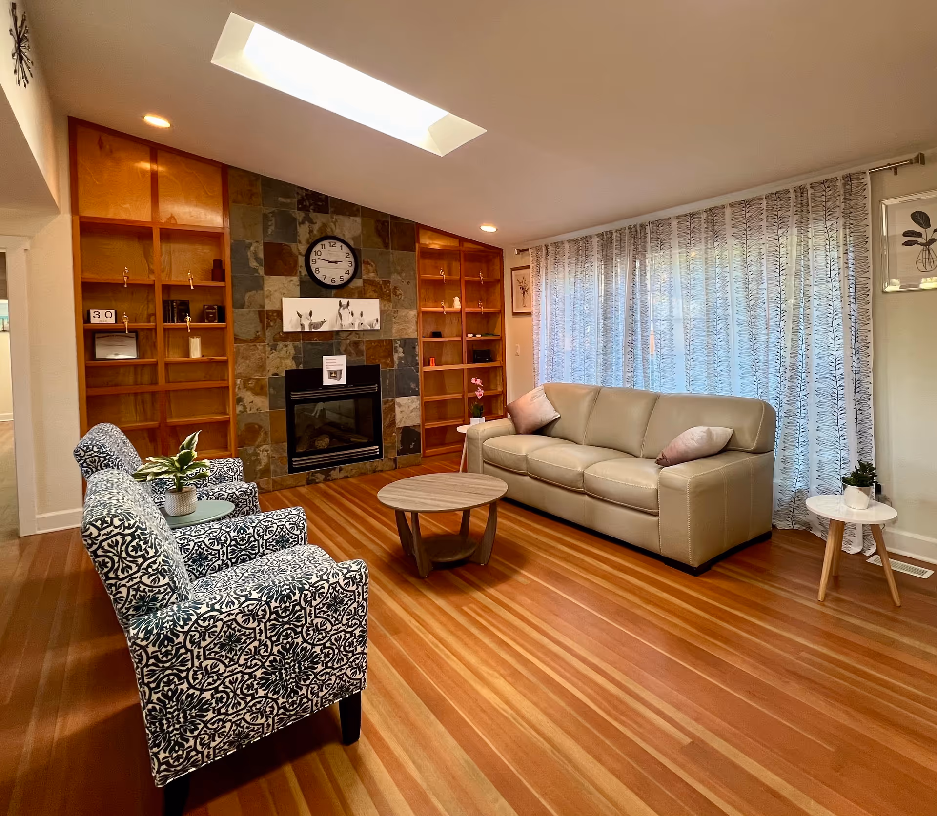 Cozy senior living lounge featuring a stone fireplace, wooden shelves, sofa, chairs, and curtains.