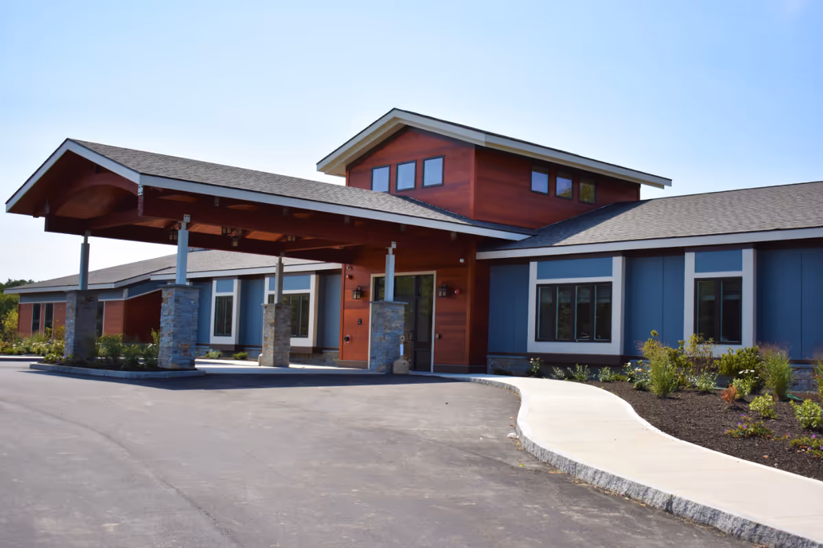 Exterior view of a modern senior living facility building with a covered entrance supported by stone pillars, surrounded by landscaped plants and a paved driveway under a clear blue sky.