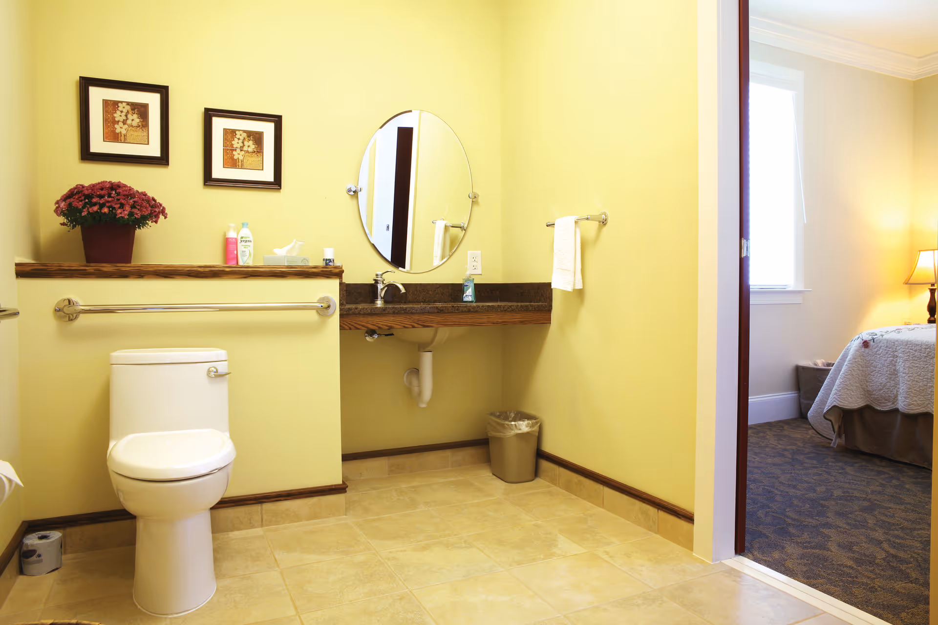 A clean, accessible bathroom with a toilet, wall-mounted grab bars, a vanity with a round mirror and toiletries, and an open doorway into a bedroom.