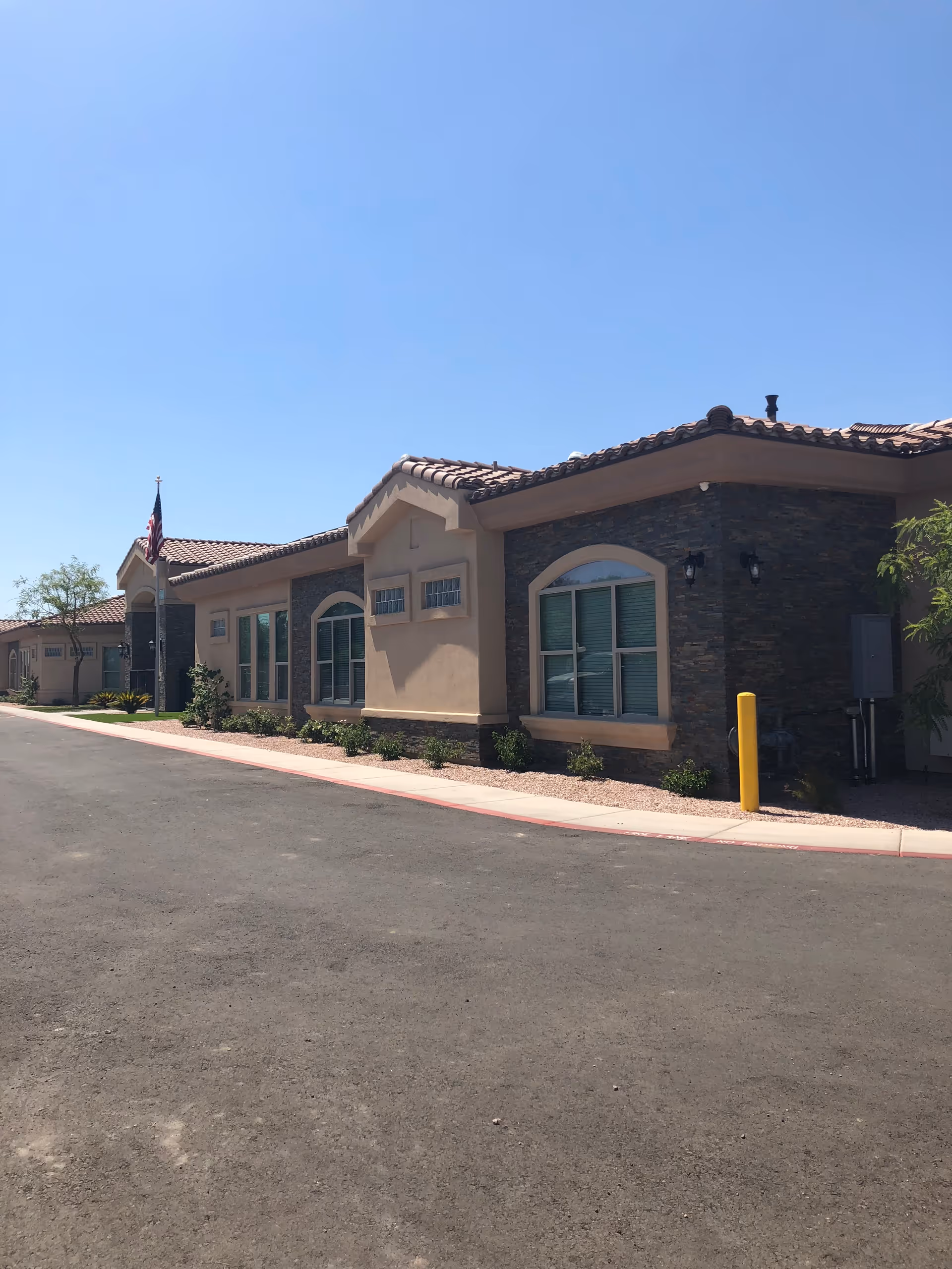 Exterior view of a single-story building with beige stucco walls, stone accents, and a tiled roof under a clear blue sky. There are several windows with white frames and an American flag near the entrance. The building is surrounded by a paved driveway and some landscaping with small bushes and trees.