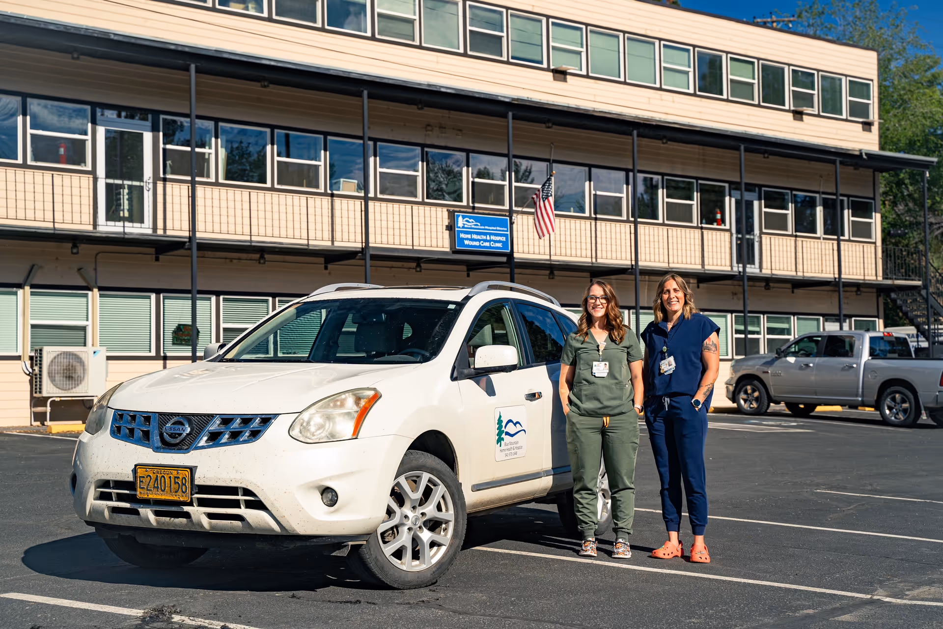 Two healthcare workers stand beside a white SUV in front of Blue Mountain Hospice Outpatient Services.