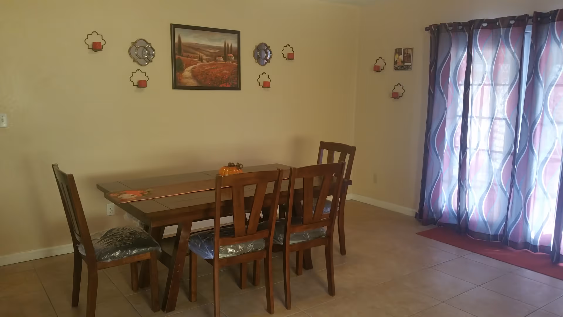 Dining room with a wooden table and six chairs, wall decorations, and patterned curtains over a sliding glass door.