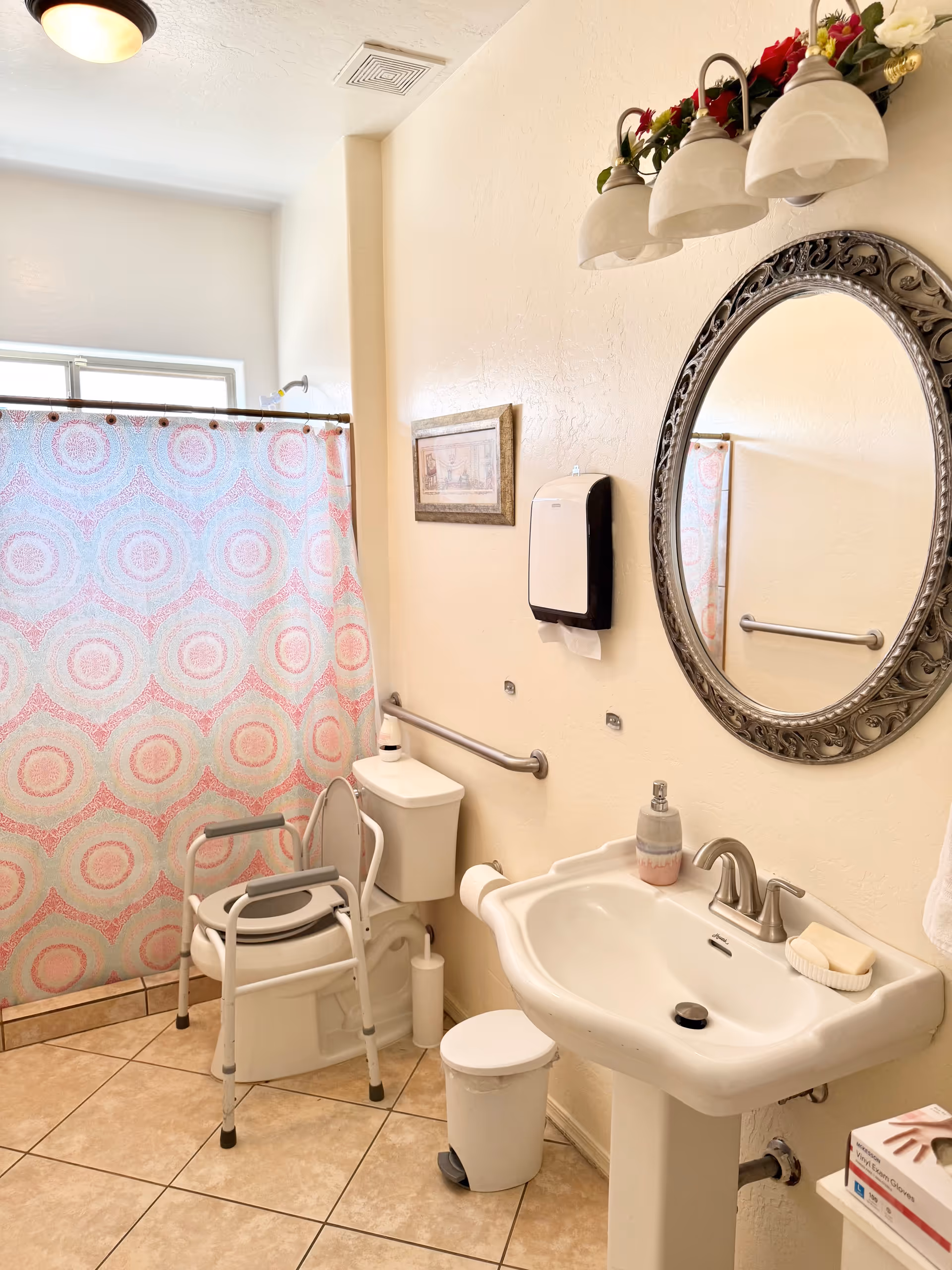 Accessible bathroom with pink patterned shower curtain, toilet, sink, mirror, and mobility chair.