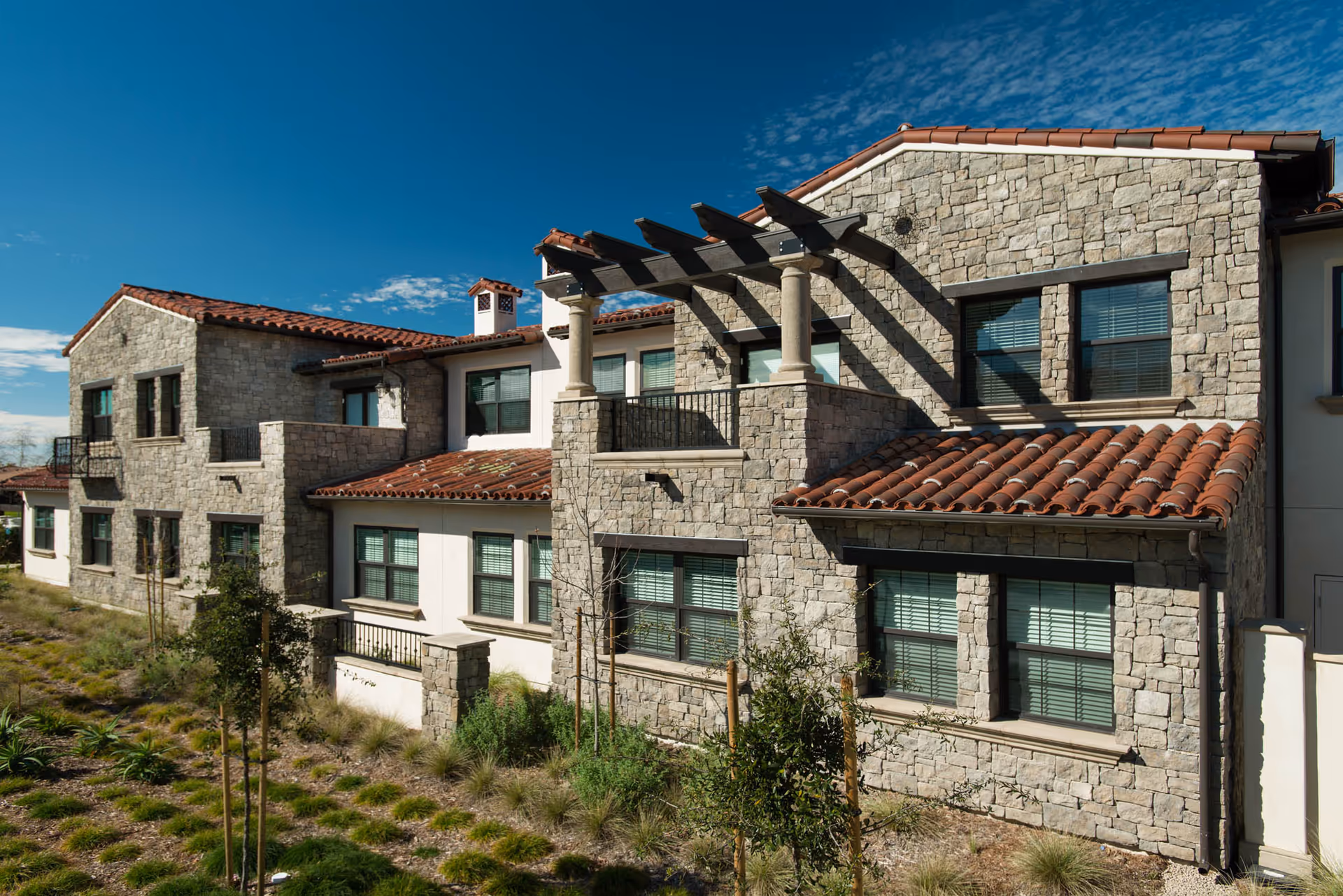Stone-clad Mediterranean-style senior living building with terracotta roof tiles, multiple windows, a balcony pergola, and landscaped grounds.