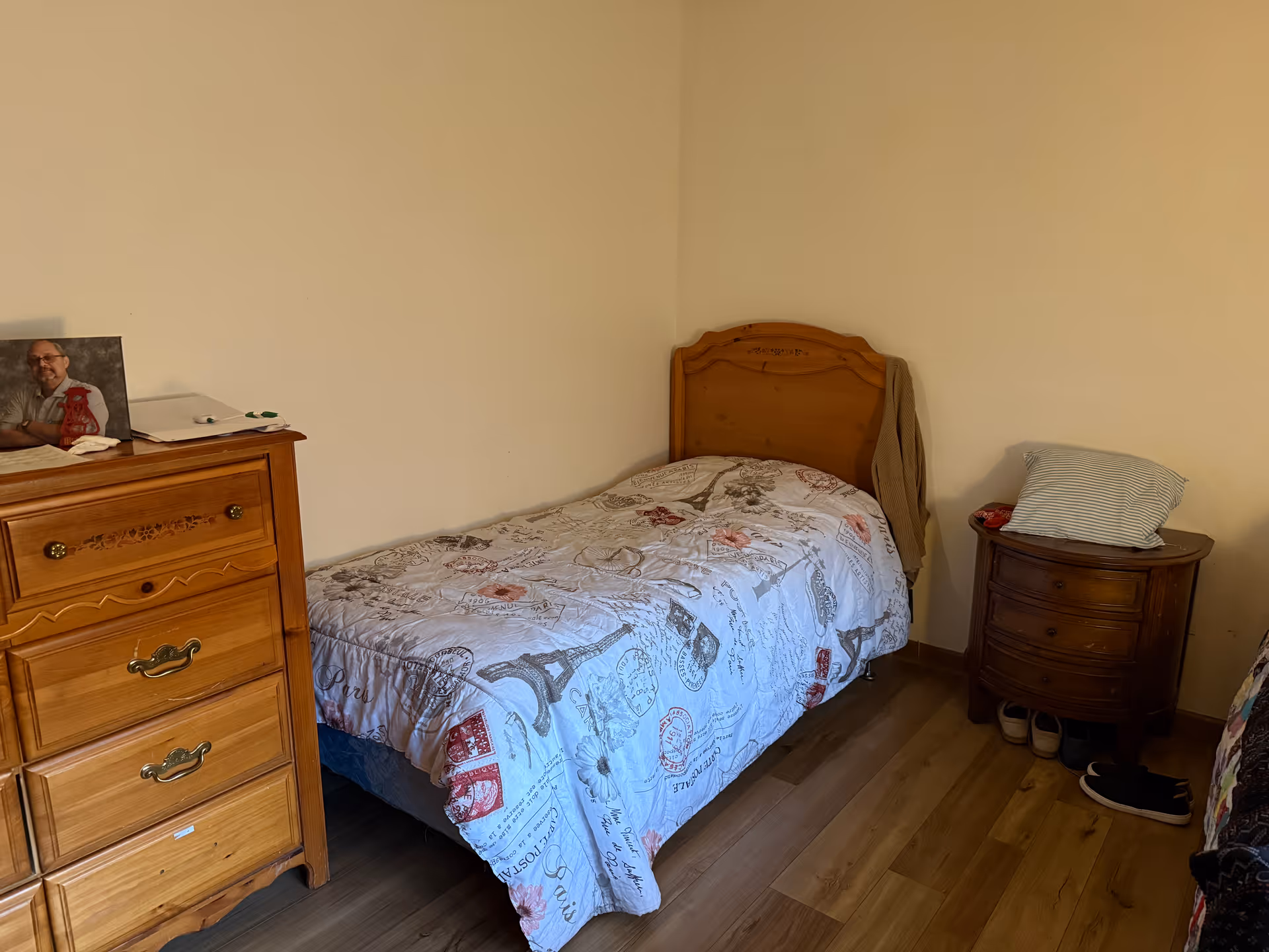 A small bedroom with a single bed, quilted cover, wooden dresser, and nightstand.