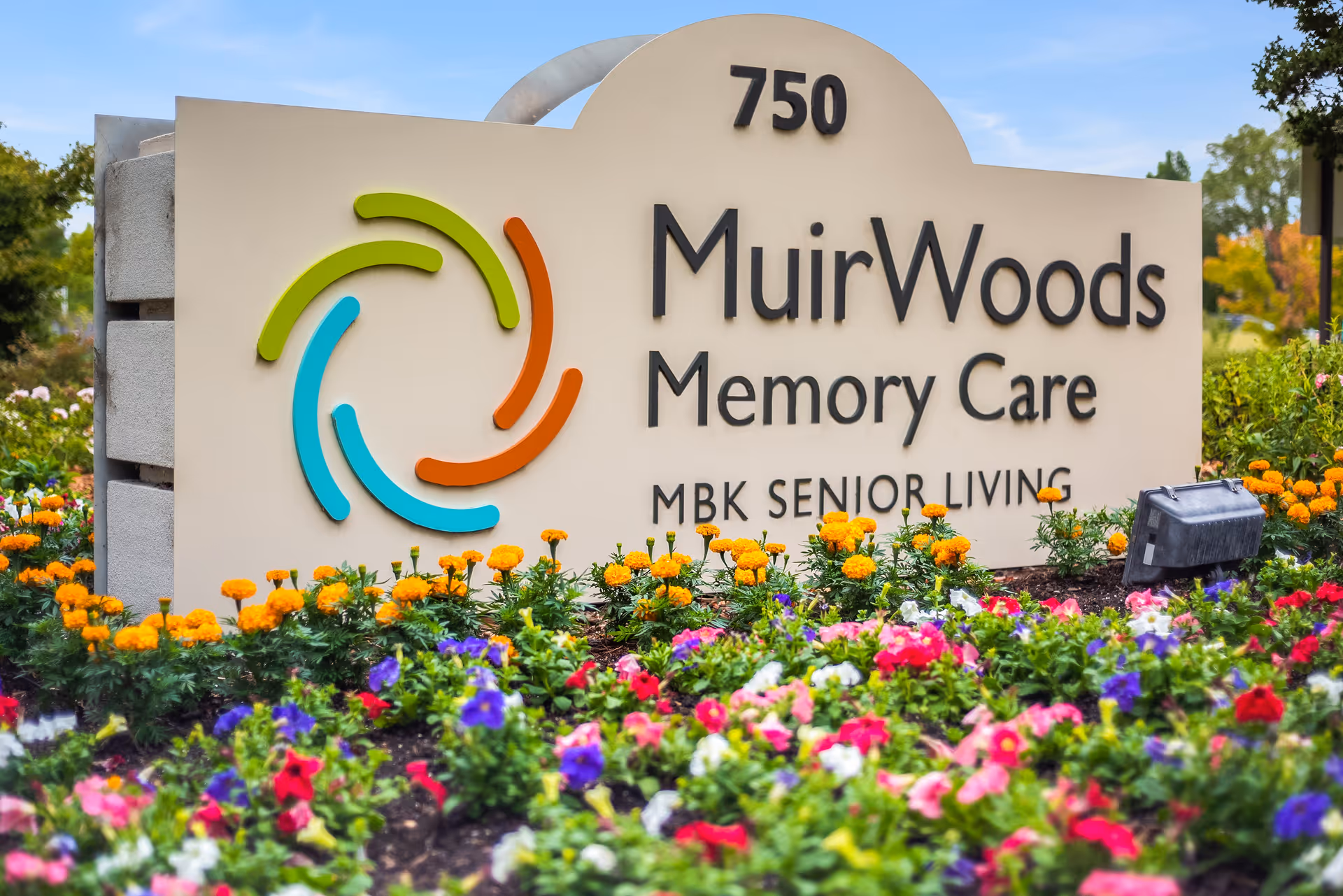 A large beige sign with the address number 750 and the text 'MuirWoods Memory Care MBK Senior Living' surrounded by colorful flowers and greenery under a clear blue sky.